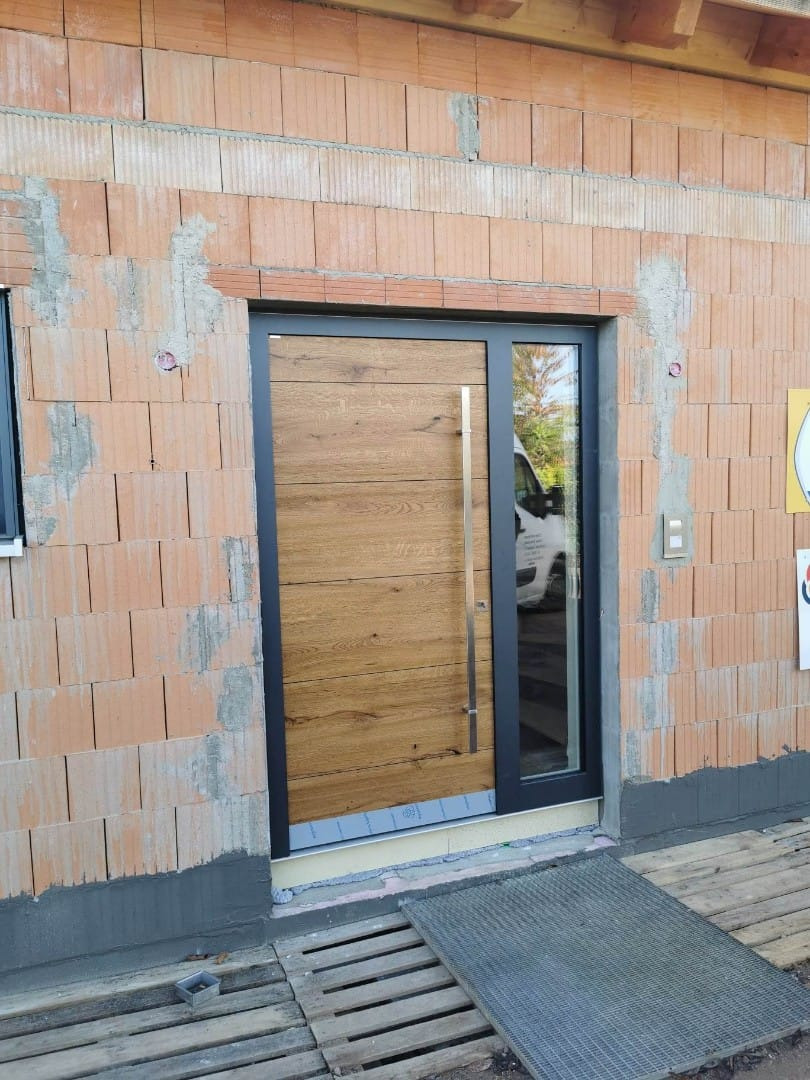 Nowoczesne drzwi wejściowe z drewnianym panelem i czarną ramą, widoczne na tle muru z czerwonej cegły w trakcie budowy.