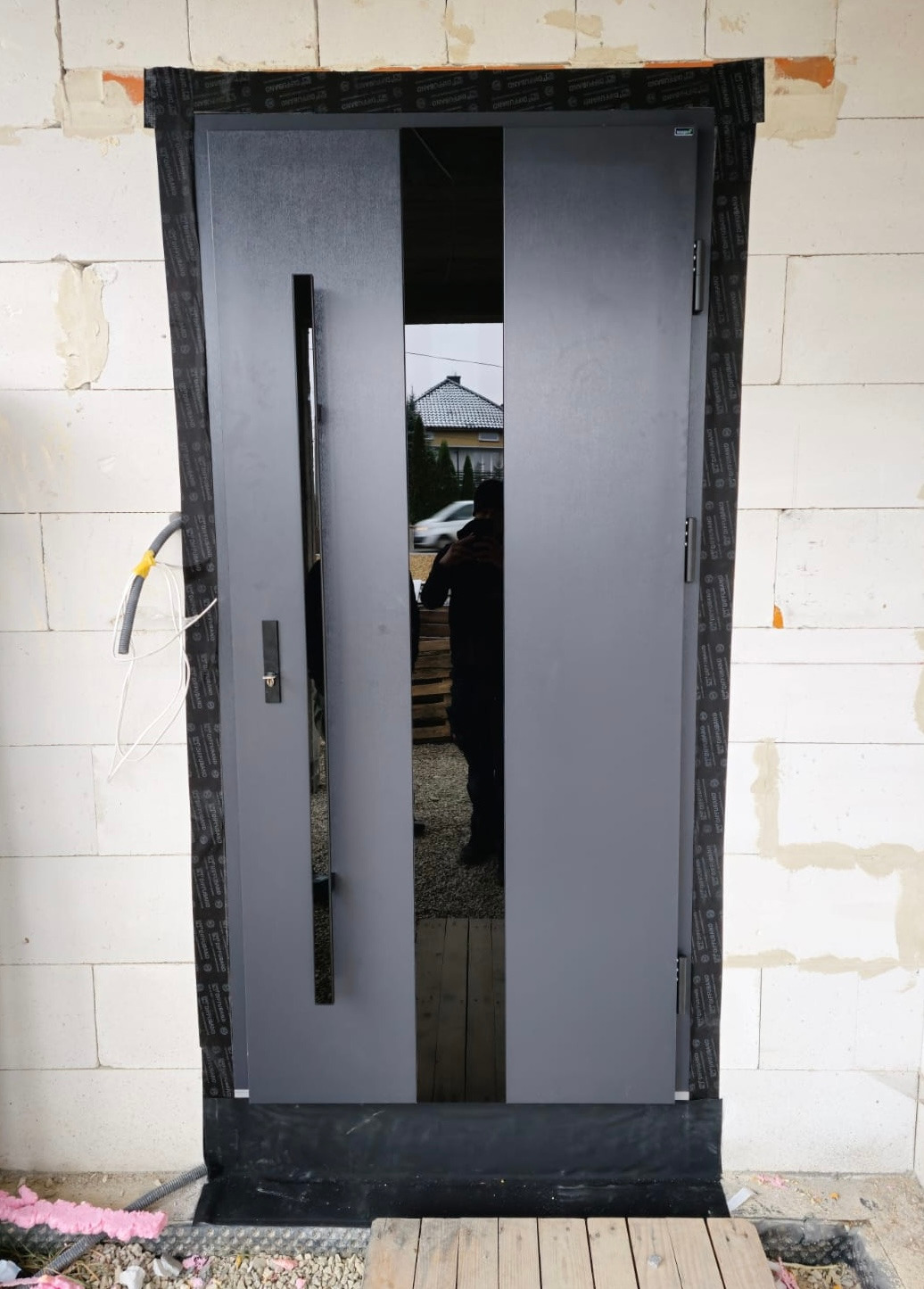 Szare, nowoczesne drzwi wejściowe z czarnym, pionowym przeszkleniem, zamontowane w surowym murze z bloczków, widoczne zabezpieczenia przeciwwilgociowe i pianka montażowa.