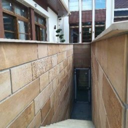 Katarzyna Chmiel Reparo - Zewnętrzne schody prowadzące do piwnicy budynku z elewacją z piaskowca, widoczne okna z brązowymi ramami i fragment dachu.