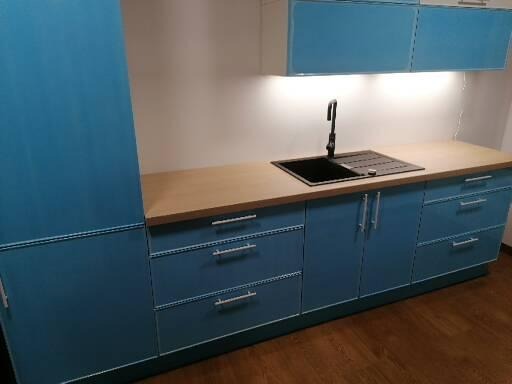 Niebieska zabudowa kuchenna z drewnianym blatem, czarnym zlewem i baterią, oświetlona od góry. Widoczne szafki wiszące i dolne z szufladami i uchwytami.