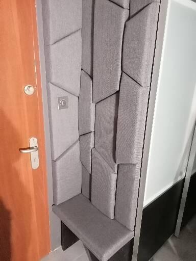 Szara tapicerowana ściana z geometrycznym wzorem 3D i siedziskiem przy pomarańczowych drzwiach z klamką.