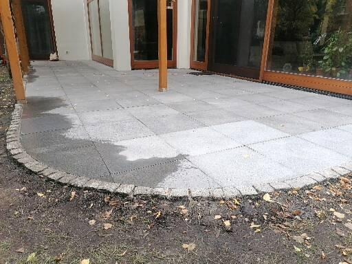 Wyłożony szarymi płytami taras z obramowaniem z kostki granitowej, przylegający do budynku z dużymi oknami tarasowymi, widoczne ślady wilgoci na powierzchni.