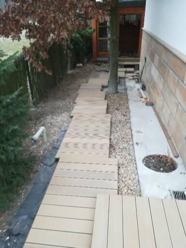 Ułożona drewniana ścieżka z desek prowadząca przez ogród, obok ściany budynku wykończonej płytkami imitującymi kamień, z widocznym drzewem rosnącym pomiędzy deskami.