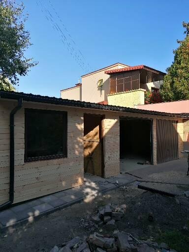 Nowo wybudowany, drewniany garaż z czarną stolarką okienną i bramą garażową z brązowych paneli, widoczny na tle domu z beżową elewacją i przeszklonym balkonem.