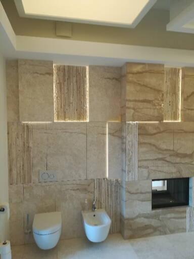 Elegancka łazienka wykończona trawertynem z podświetleniem LED w pionowych panelach, wisząca miska WC i bidet w minimalistycznym stylu.