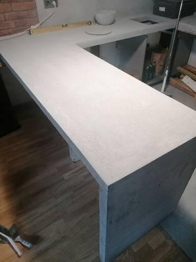 Szara, betonowa umywalka w trakcie montażu w łazience, widoczny fragment ściany z cegły i drewniana podłoga.