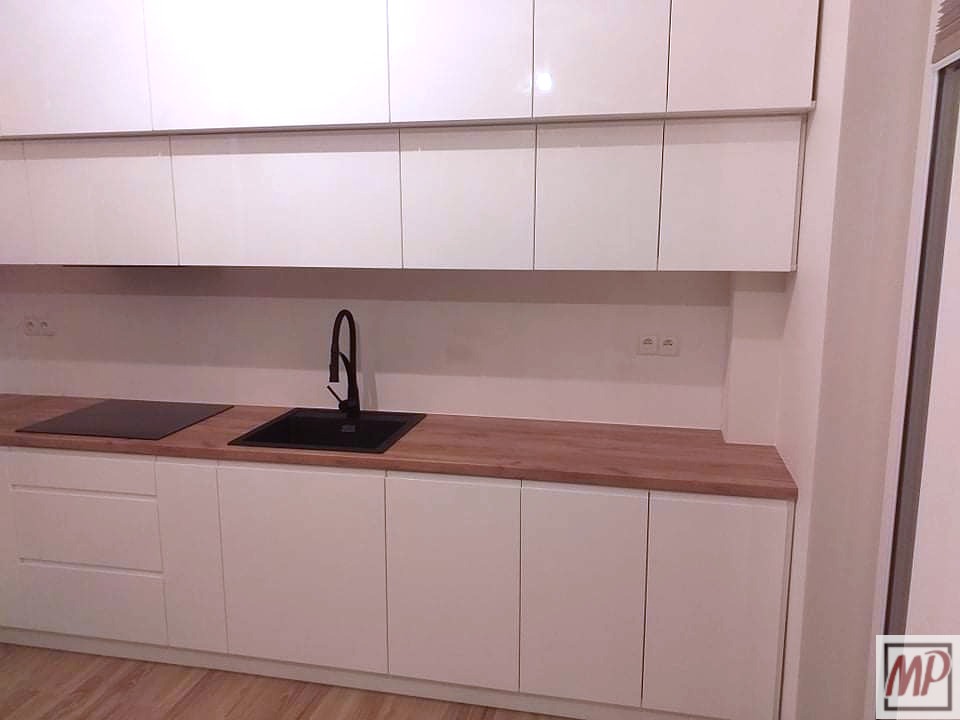 Nowoczesna, biała zabudowa kuchenna z drewnianym blatem, czarnym zlewem i minimalistyczną baterią, widoczna płyta indukcyjna.