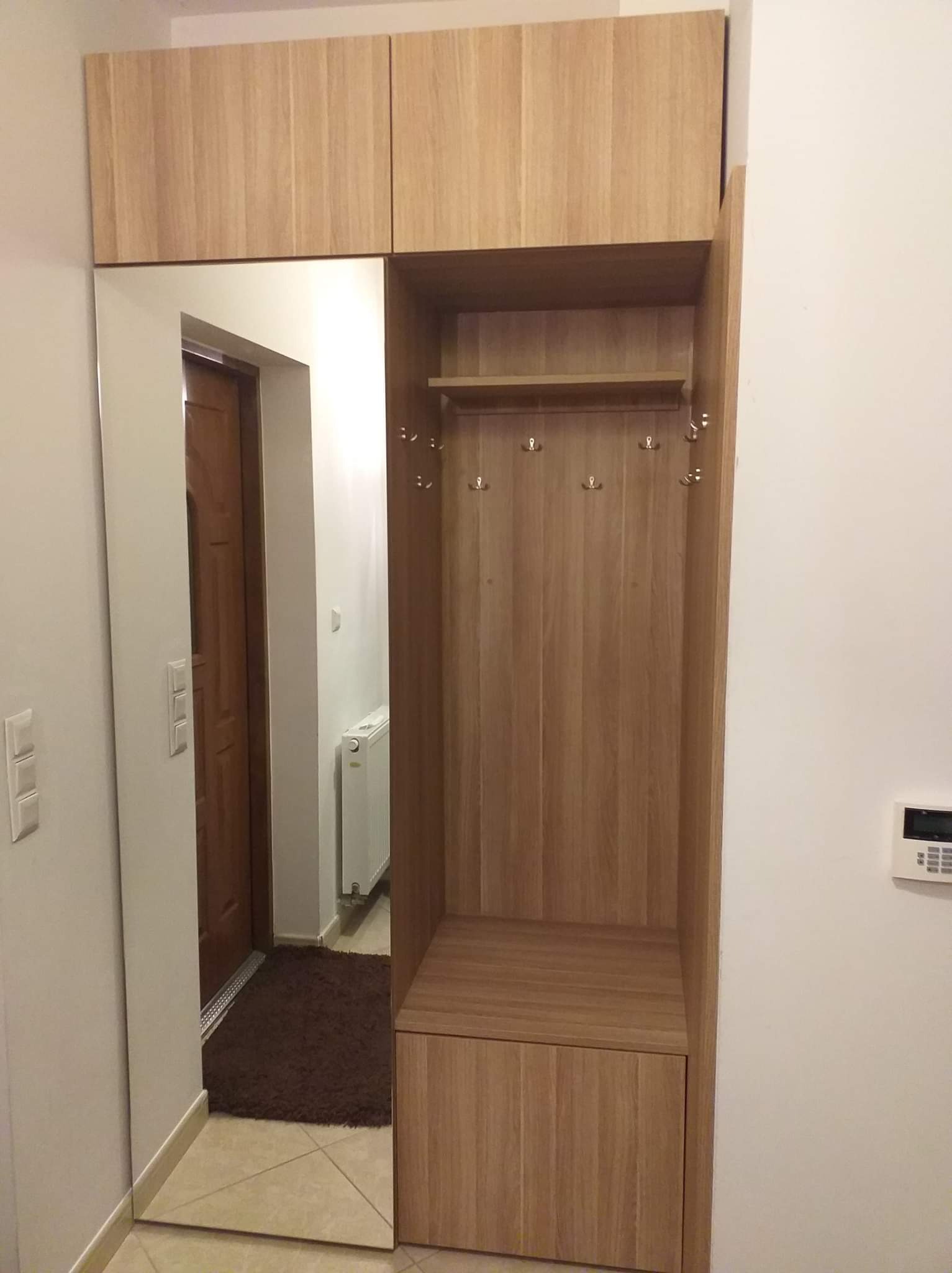 Drewniana zabudowa przedpokoju z lustrem i wieszakami, jasne drewno, widok na drzwi wejściowe i brązowy dywanik.