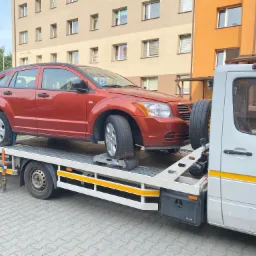 Czerwony samochód osobowy, Dodge Caliber, zabezpieczony na platformie białej lawety na tle bloku mieszkalnego.