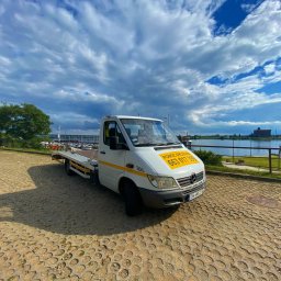 Pomoc Drogowa Wrocław 24/7 D.R. - Biała laweta Mercedes-Benz z żółtym napisem 'POMOC DROGOWA 663 017 398' na tle błękitnego nieba z obłokami, zaparkowana na betonowej nawierzchni w pobliżu rzeki i mostu.