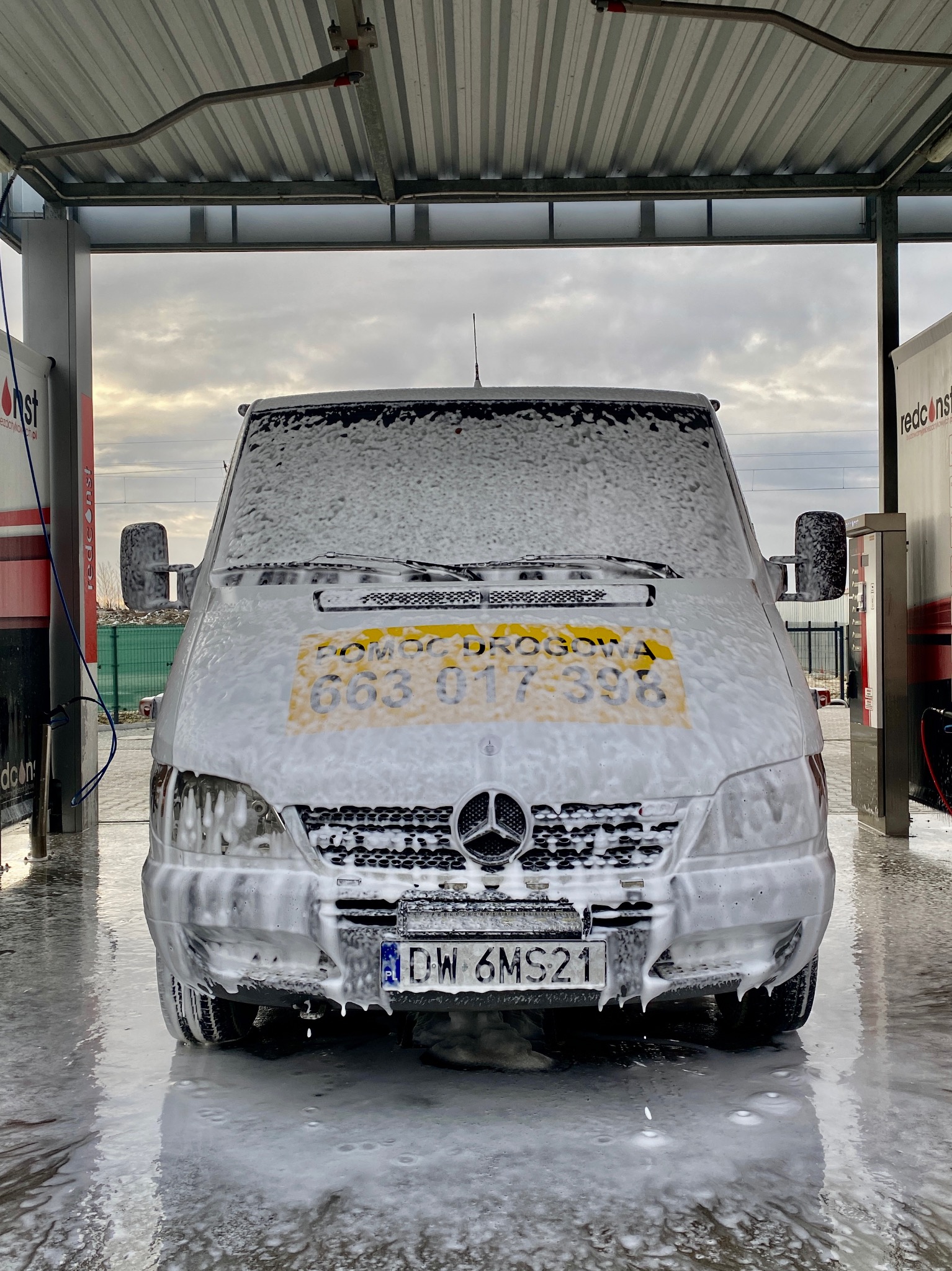 Przód białego busa Mercedes Sprinter obficie pokrytego pianą w myjni samochodowej we Wrocławiu. Na żółtym tle widoczny napis POMOC DROGOWA i numer telefonu.