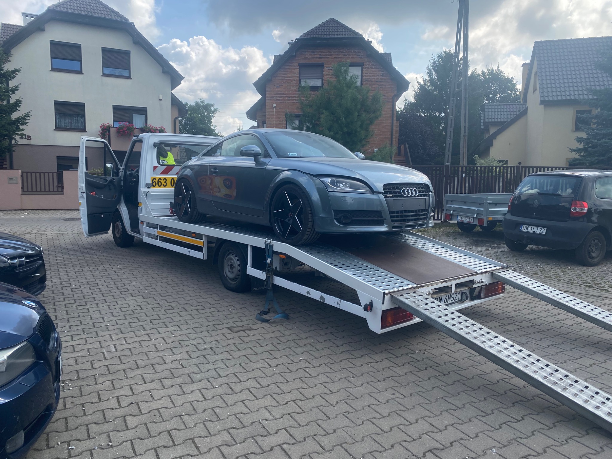 Szara Audi TT na białej lawecie z otwartymi drzwiami kabiny, zaparkowana na brukowanej powierzchni przed domami.