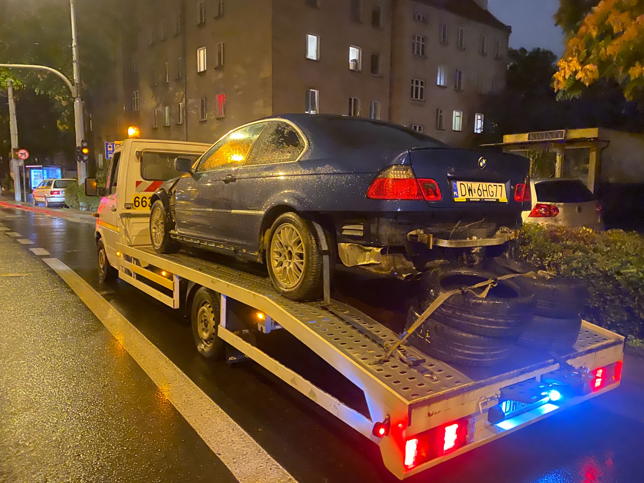 Nocne ujęcie uszkodzonego, granatowego BMW serii 3 na platformie lawety, z widocznymi zapasowymi oponami zabezpieczonymi pasami, na mokrej ulicy w tle budynek mieszkalny z zapalonymi oknami.