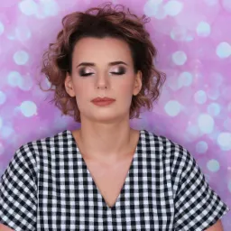 Portret kobiety z zamkniętymi oczami, prezentującej wieczorowy makijaż z błyszczącymi cieniami i matową pomadką, na tle fioletowego, rozmytego tła z efektem bokeh. Widoczne logo firmy w prawym...