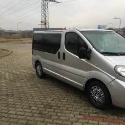 Srebrny van z przyciemnianymi szybami zaparkowany na kostce brukowej, w tle słup wysokiego napięcia i pochmurne niebo.