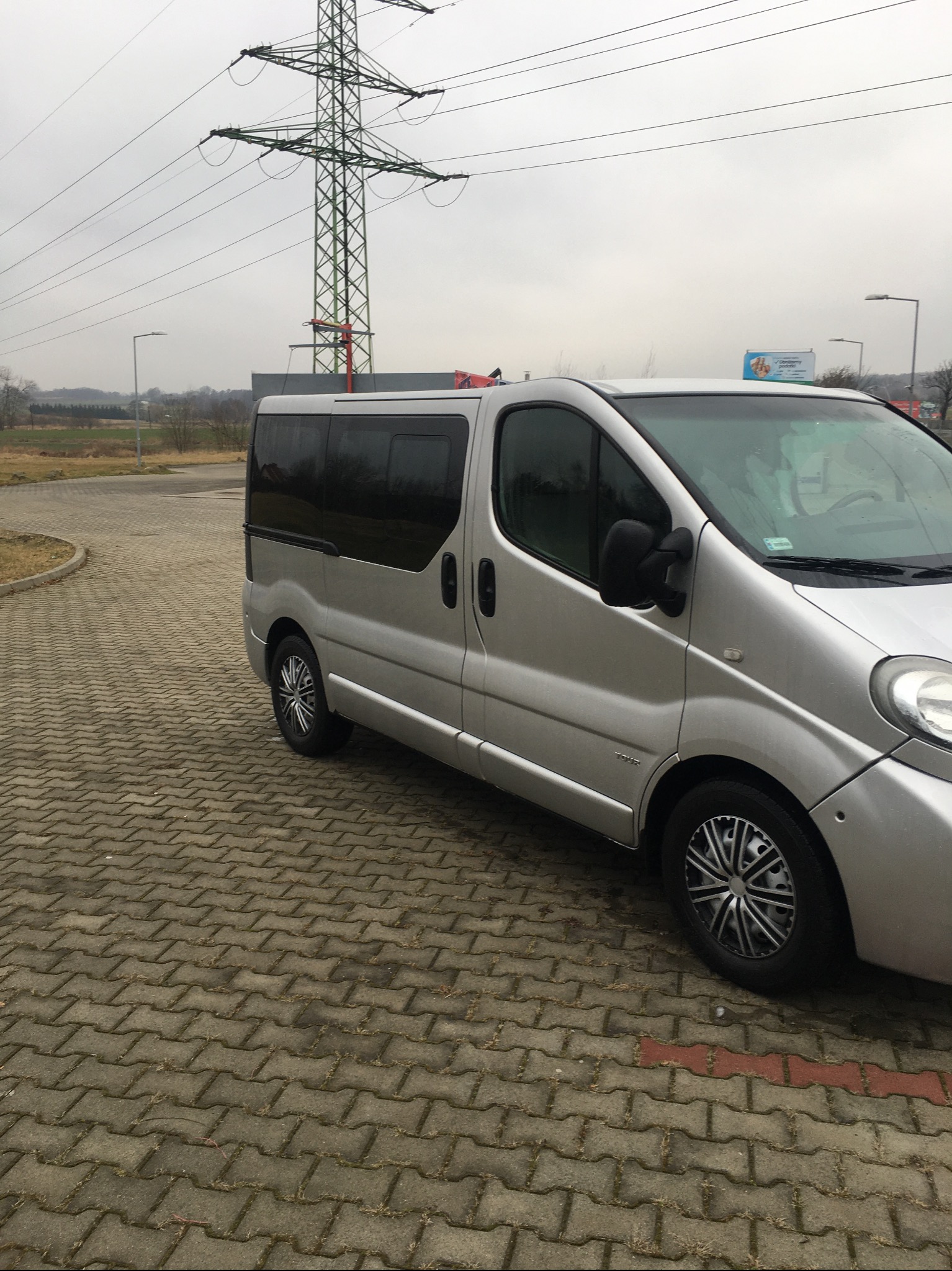Srebrny van z przyciemnianymi szybami zaparkowany na kostce brukowej, w tle słup wysokiego napięcia i pochmurne niebo.