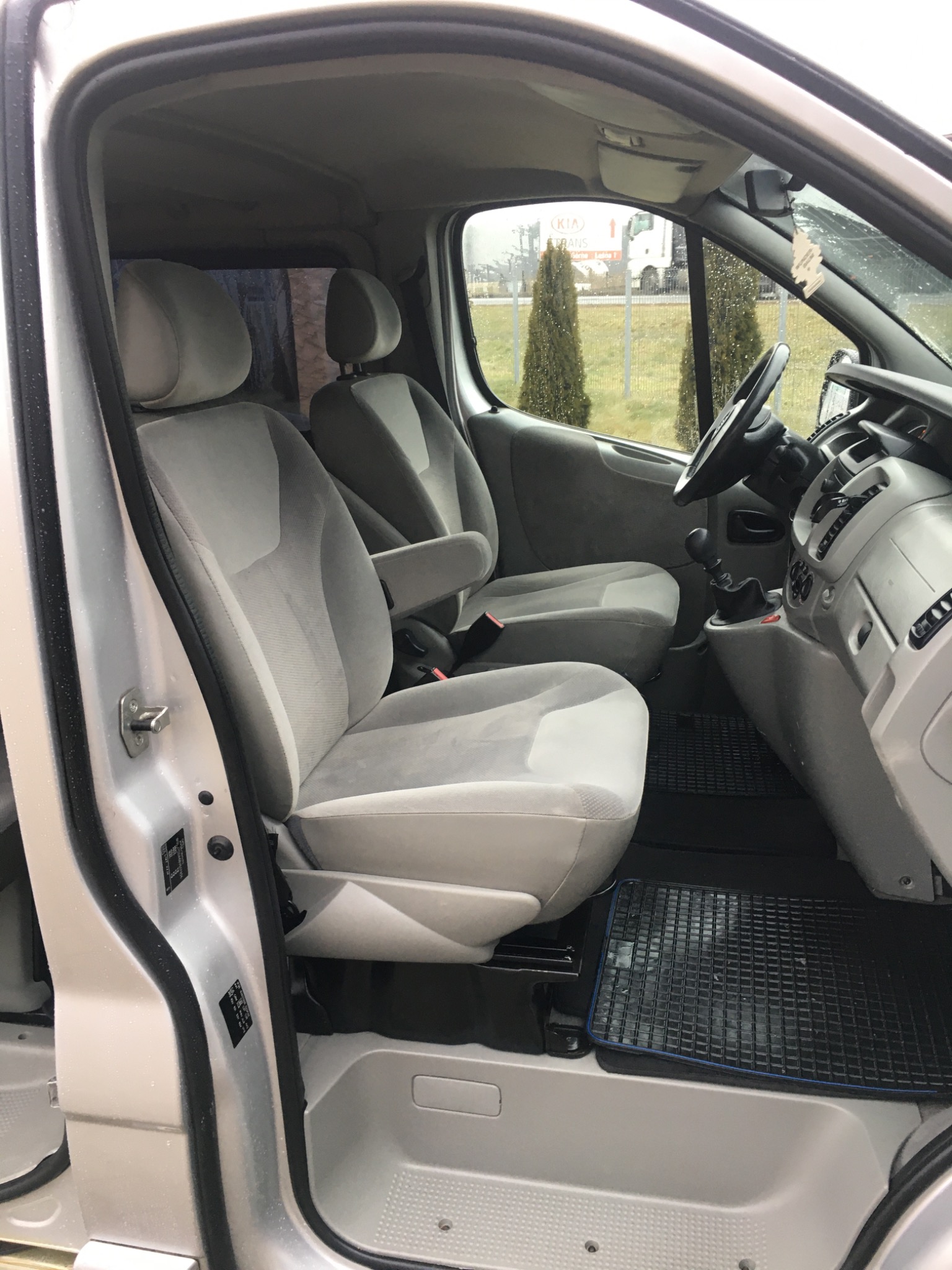 Wnętrze srebrnego busa z szarymi fotelami, widok z otwartych drzwi kierowcy, na podłodze czarna gumowa wycieraczka z niebieską obwódką.