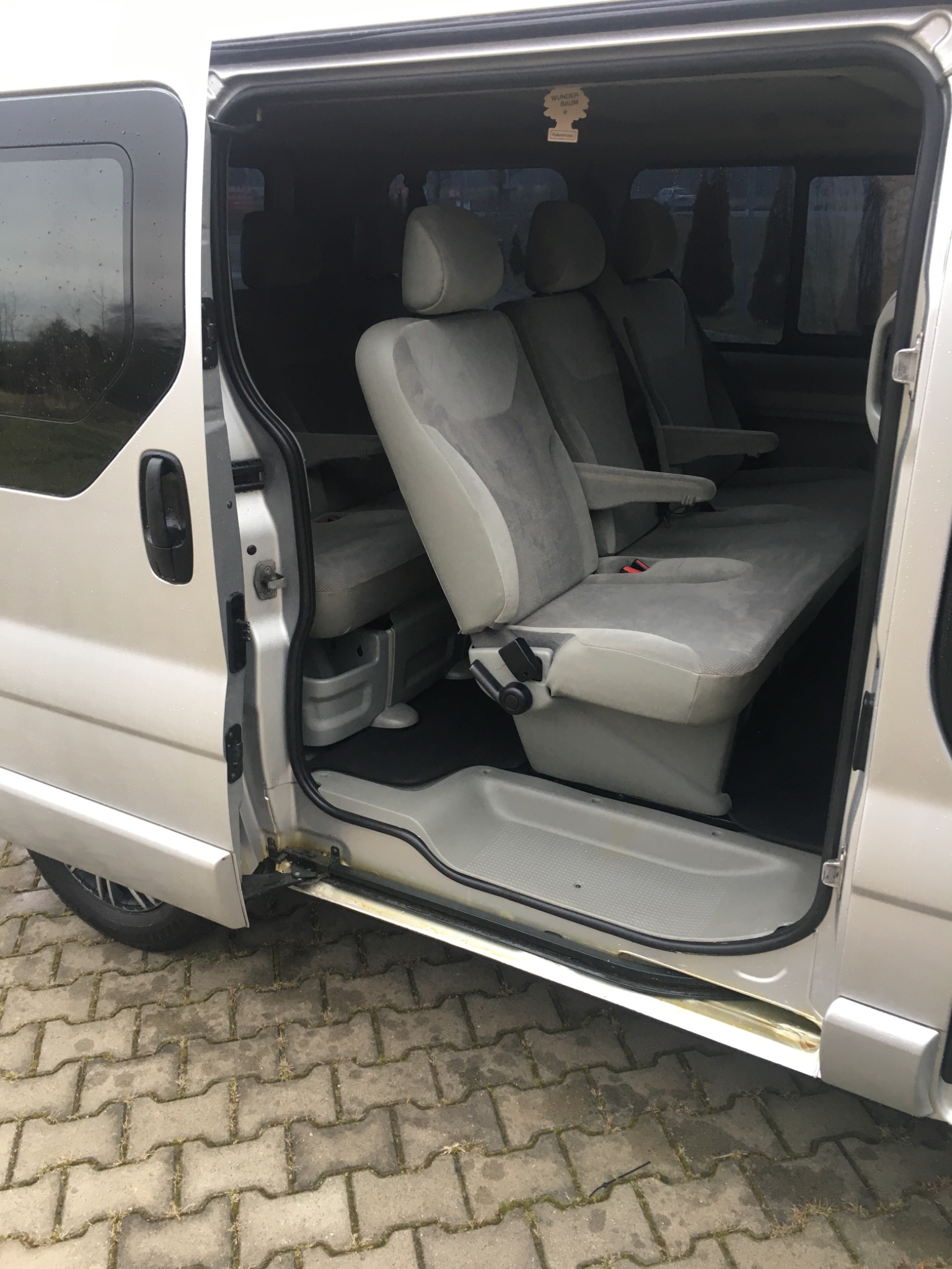 Wnętrze srebrnego busa z otwartymi drzwiami, widoczne szare welurowe fotele pasażerskie i zapach samochodowy Wunder-Baum.