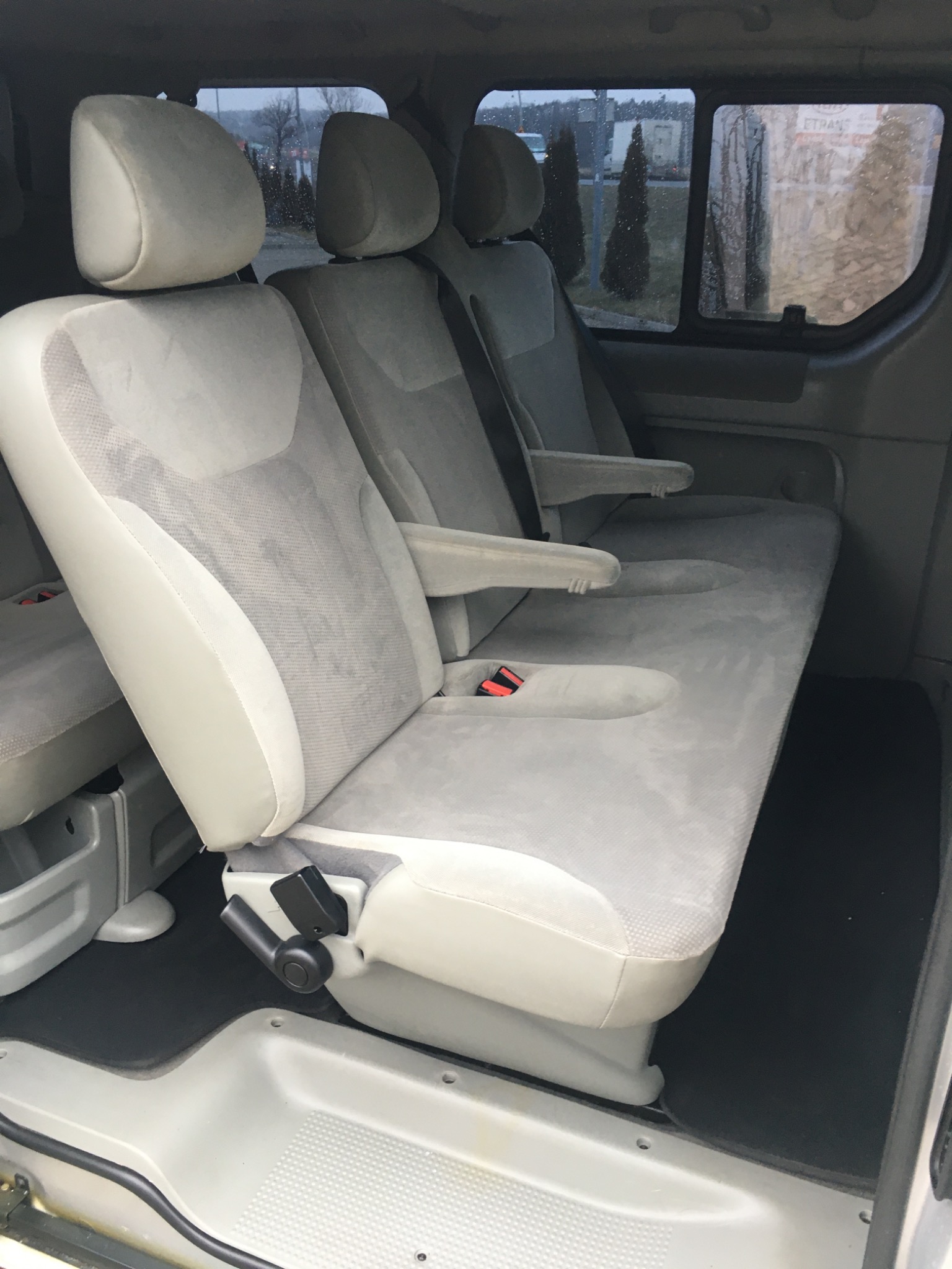 Wnętrze busa z trzema rzędami szarych foteli z podłokietnikami, widok z tyłu pojazdu, za oknem widoczny parking i drzewa.