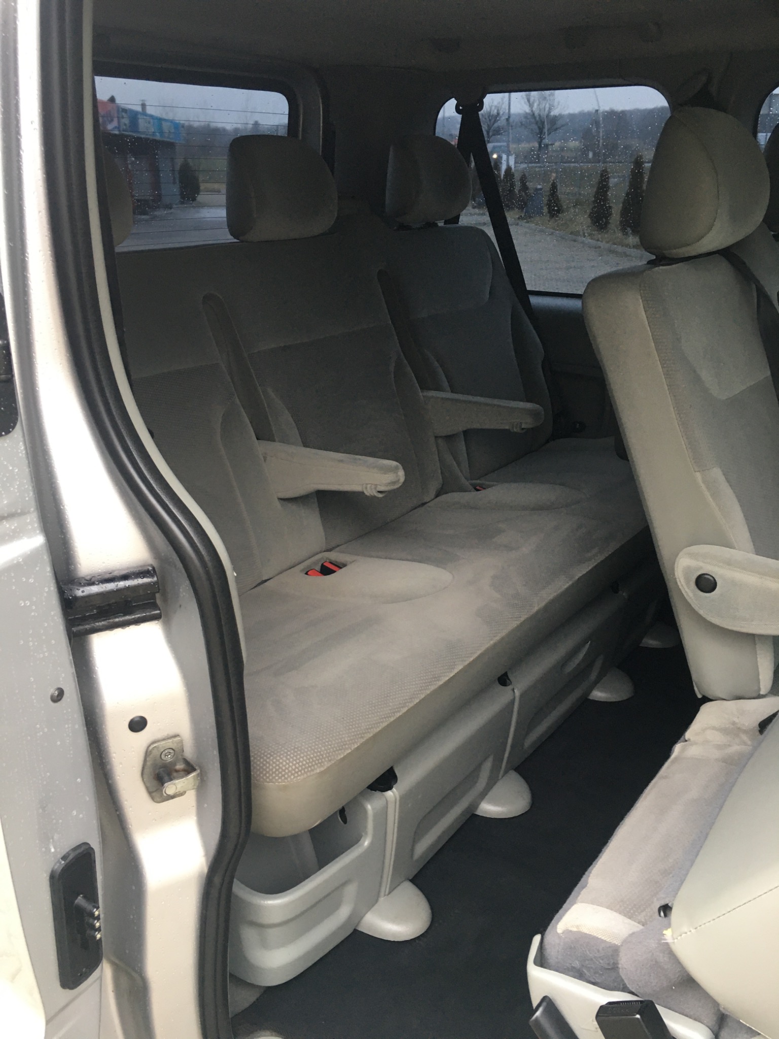 Wnętrze srebrnego vana z trzema rzędami szarych foteli z podłokietnikami, widok z otwartych drzwi bocznych na mokry parking.