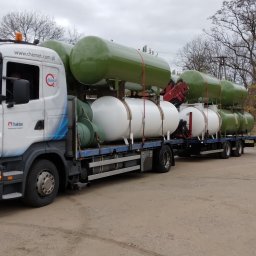 GAS-SERWIS ADAM SUCHECKI - Ciężarówka Scania przewozi na platformie zestaw białych i zielonych zbiorników gazowych, zabezpieczonych czerwonymi pasami, na tle drzew i budynku.