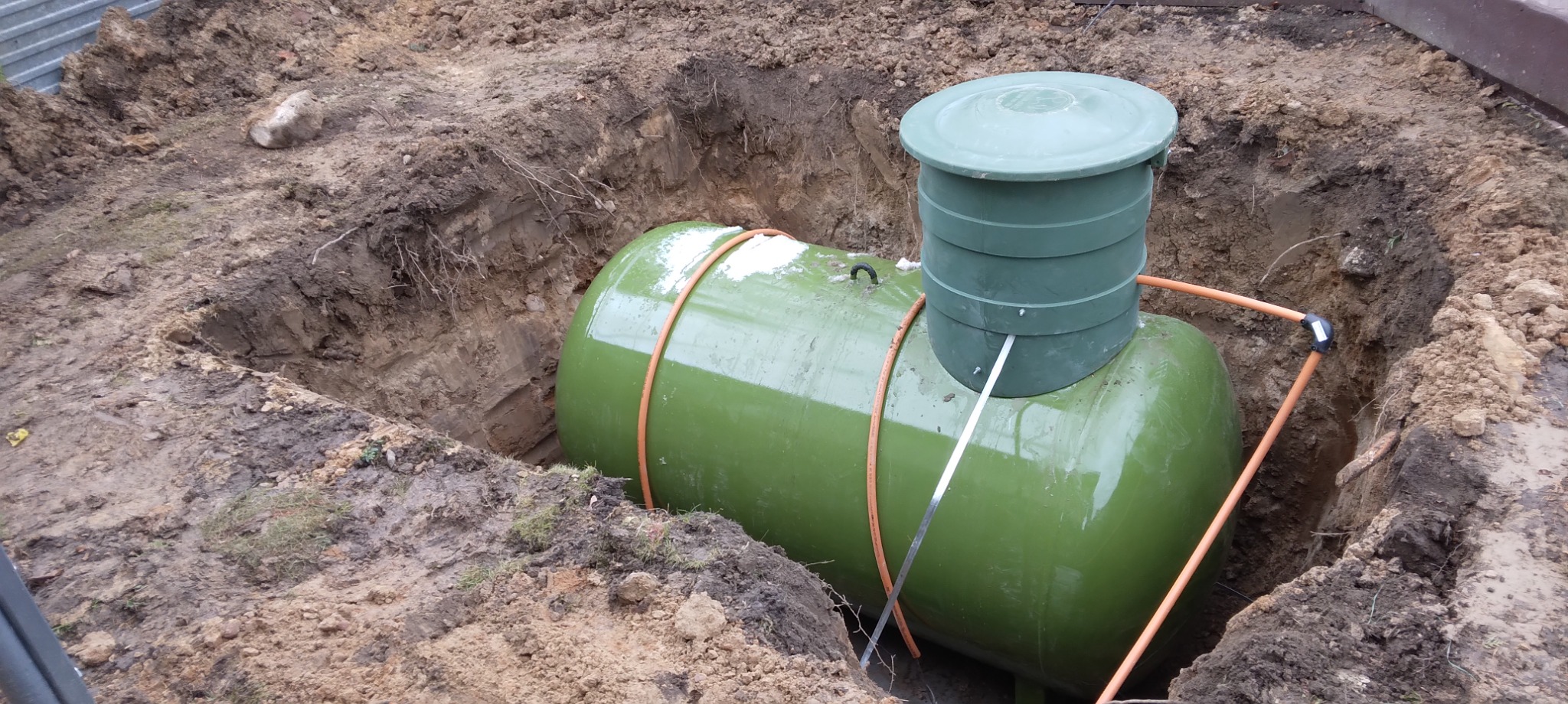 Zielony zbiornik na gaz płynny częściowo zakopany w wykopie ziemnym, z widocznymi rurami doprowadzającymi i włazem inspekcyjnym.