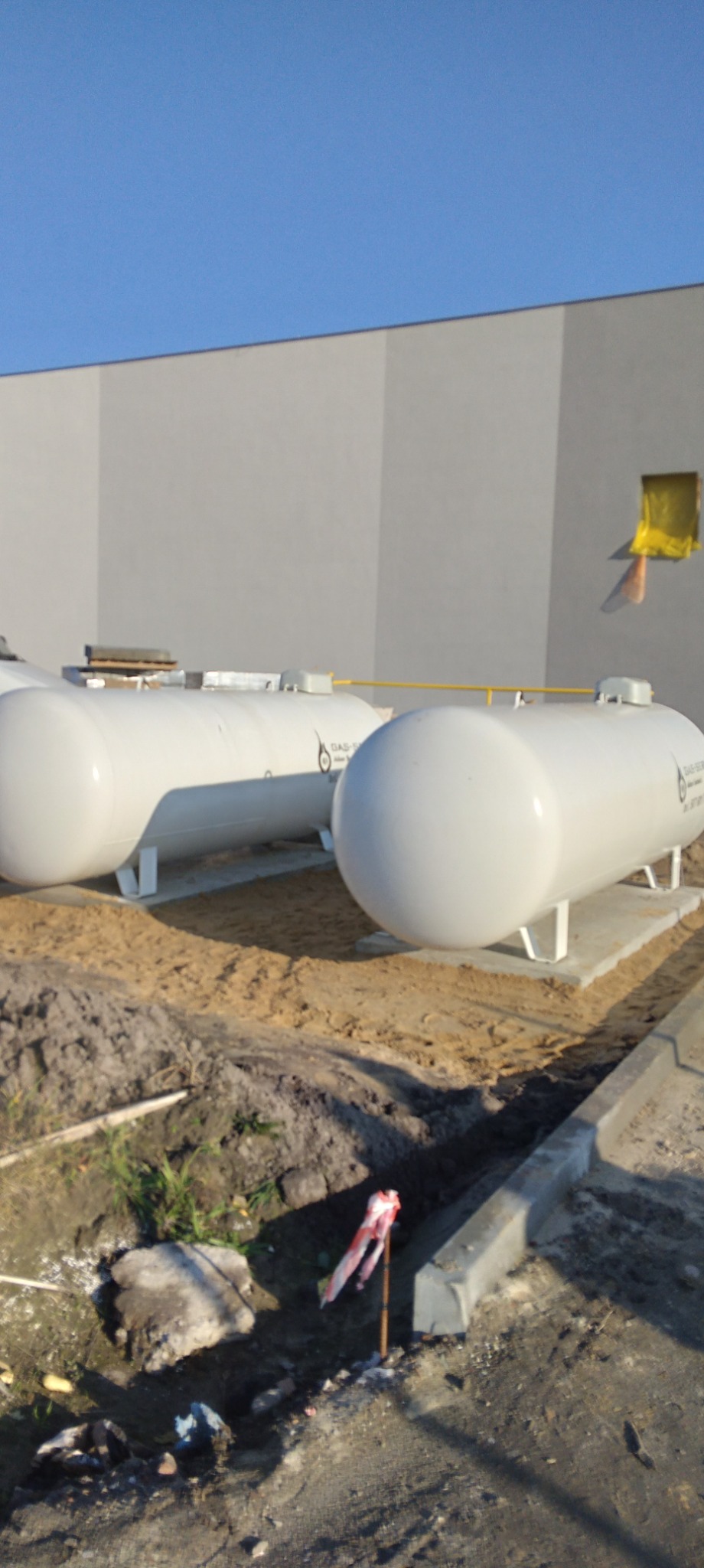 Dwa białe zbiorniki gazu na betonowych fundamentach przed szarą ścianą budynku, widoczne żółte rury łączące zbiorniki.