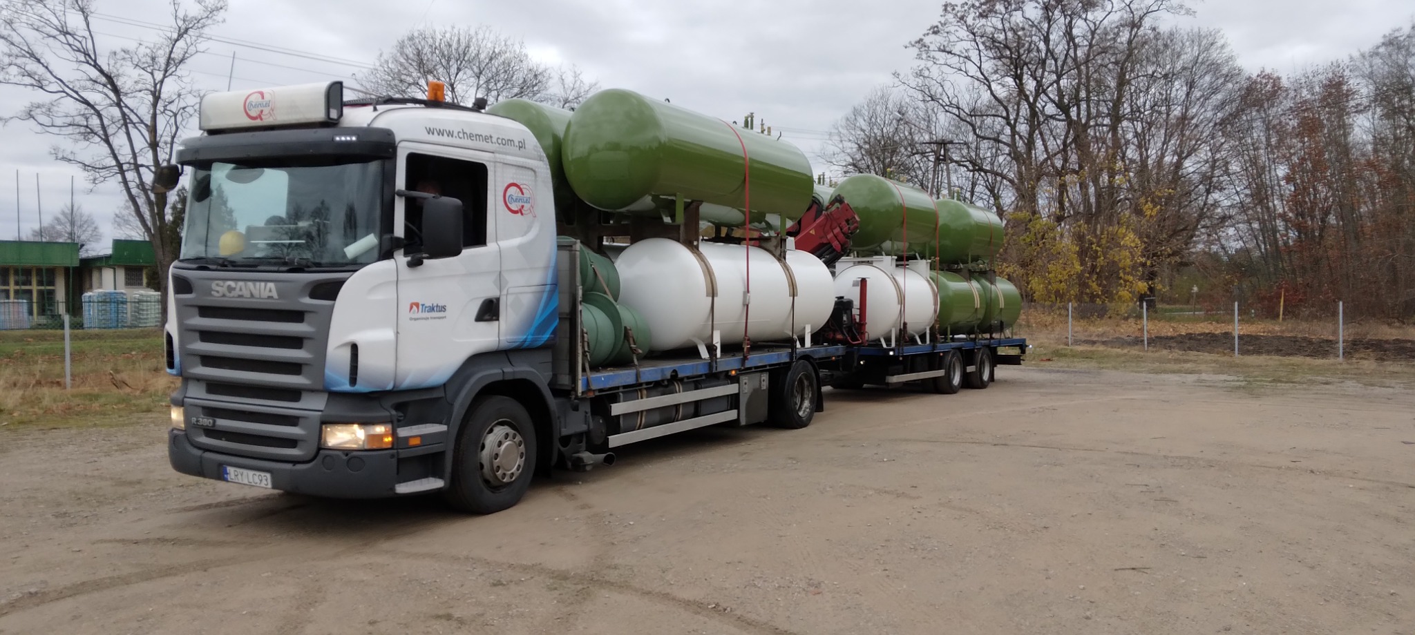 Ciężarówka Scania przewozi na platformie zestaw białych i zielonych zbiorników gazowych, zabezpieczonych czerwonymi pasami, na tle drzew i budynku.