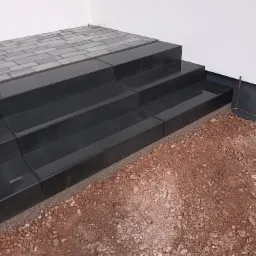 Ciemne, eleganckie schody z kostki brukowej prowadzące do budynku, otoczone ziemią. Widoczne detale łączeń i faktura materiału.