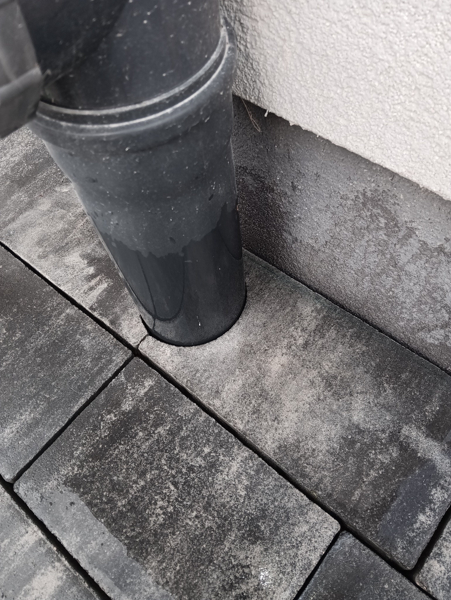 Ciemnoszara kostka brukowa ułożona przy rurze spustowej, blisko ściany budynku. Widoczne detale faktury kostki i precyzyjne dopasowanie do rury.