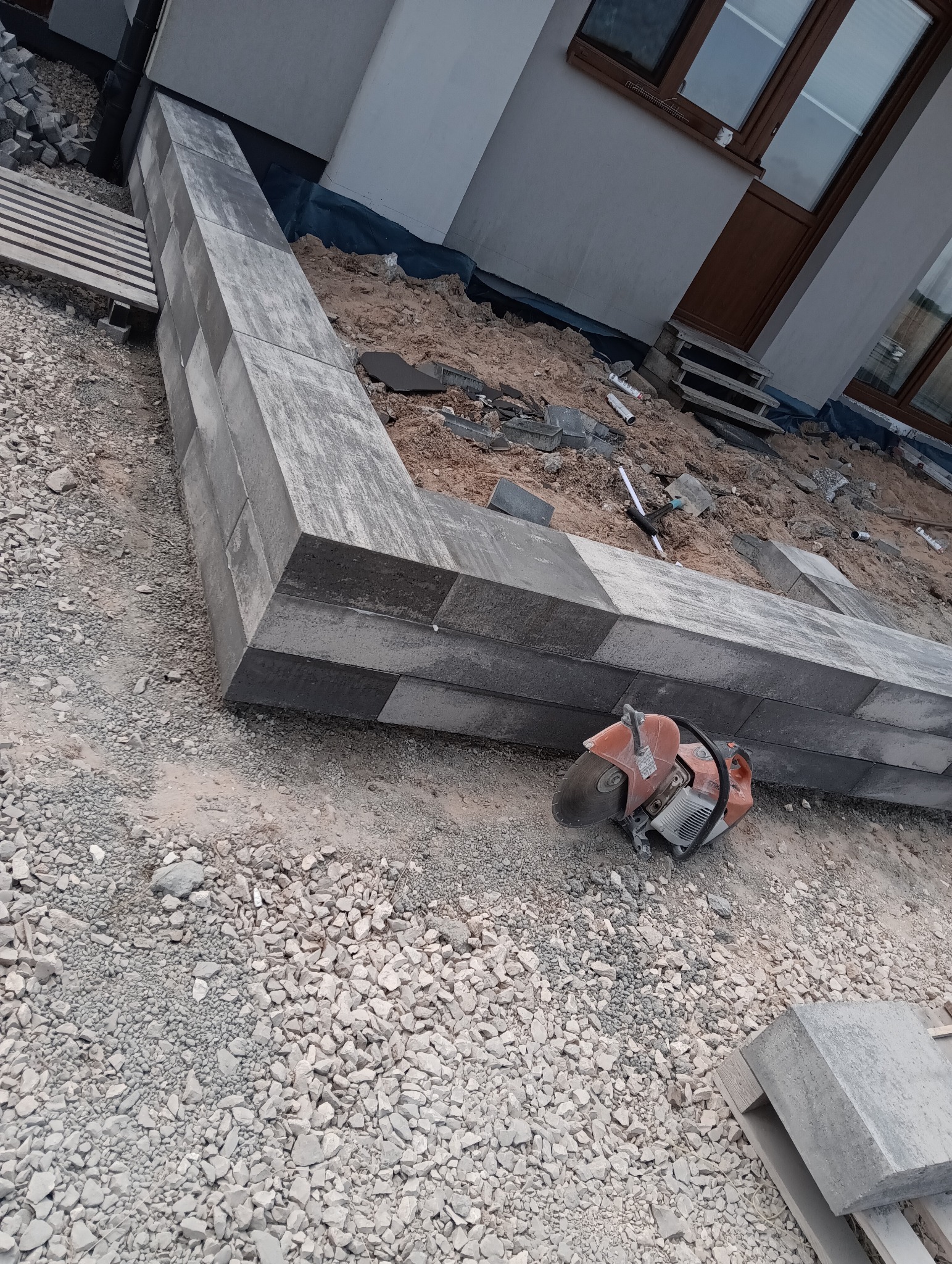 Wzniesiona obudowa z szarej kostki brukowej, piła do cięcia betonu, gruz i podłoże przed budynkiem. Etap prac brukarskich z użyciem narzędzi.