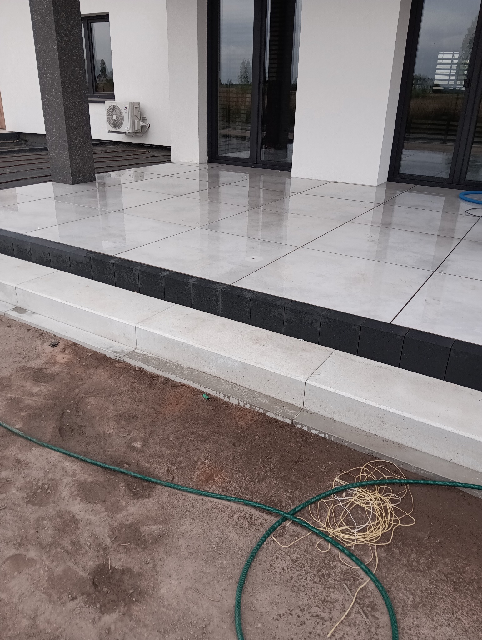 Białe, błyszczące płyty tarasowe z czarnym obrzeżem przy nowoczesnym domu. W tle widoczne okna i klimatyzator. W dole ziemia i węże.