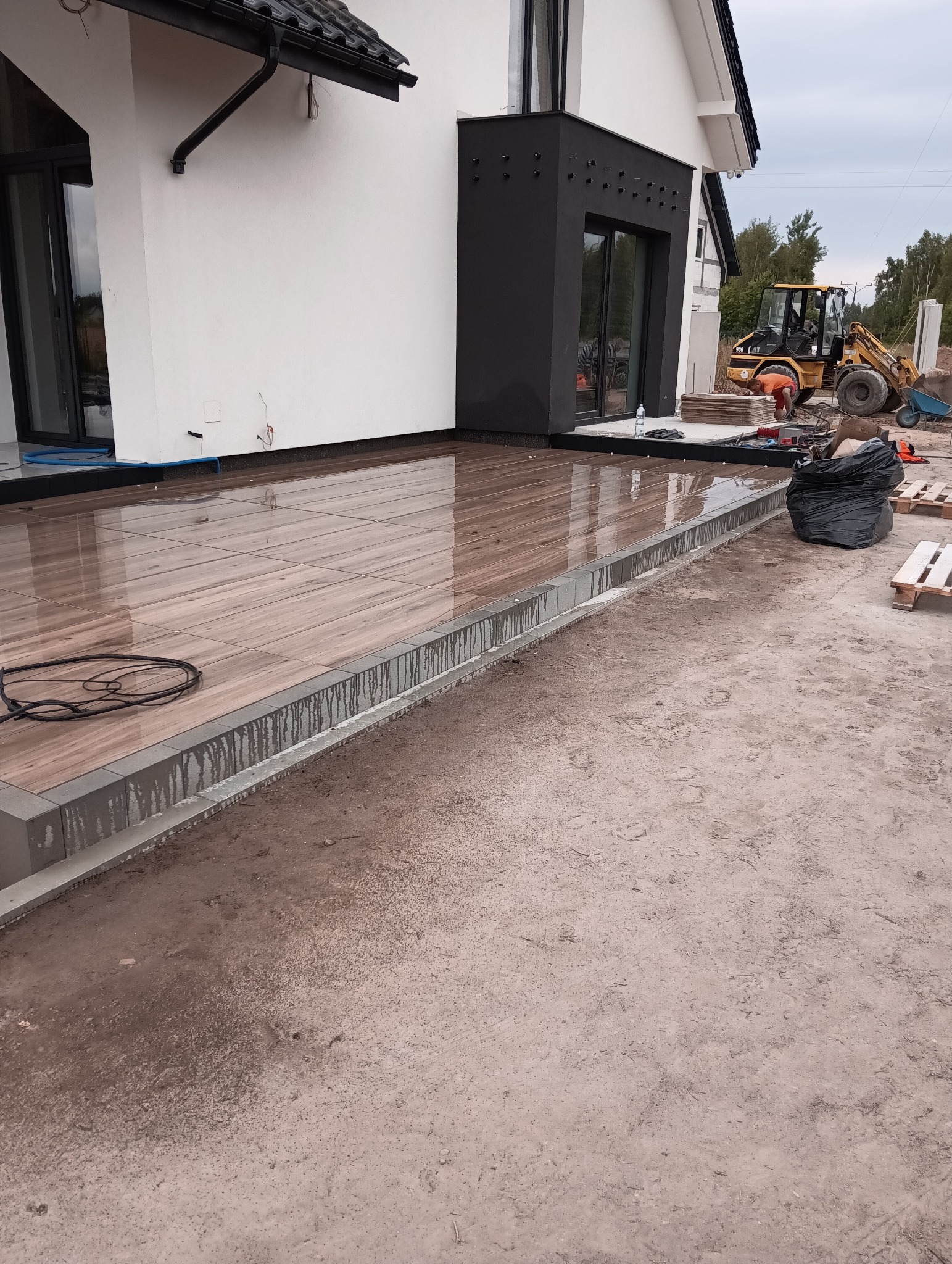 Nowoczesny taras z płytek imitujących drewno, obrzeża z szarej kostki, w tle budynek i koparka. Prace brukarskie w toku, widoczny teren przygotowany pod dalsze prace.