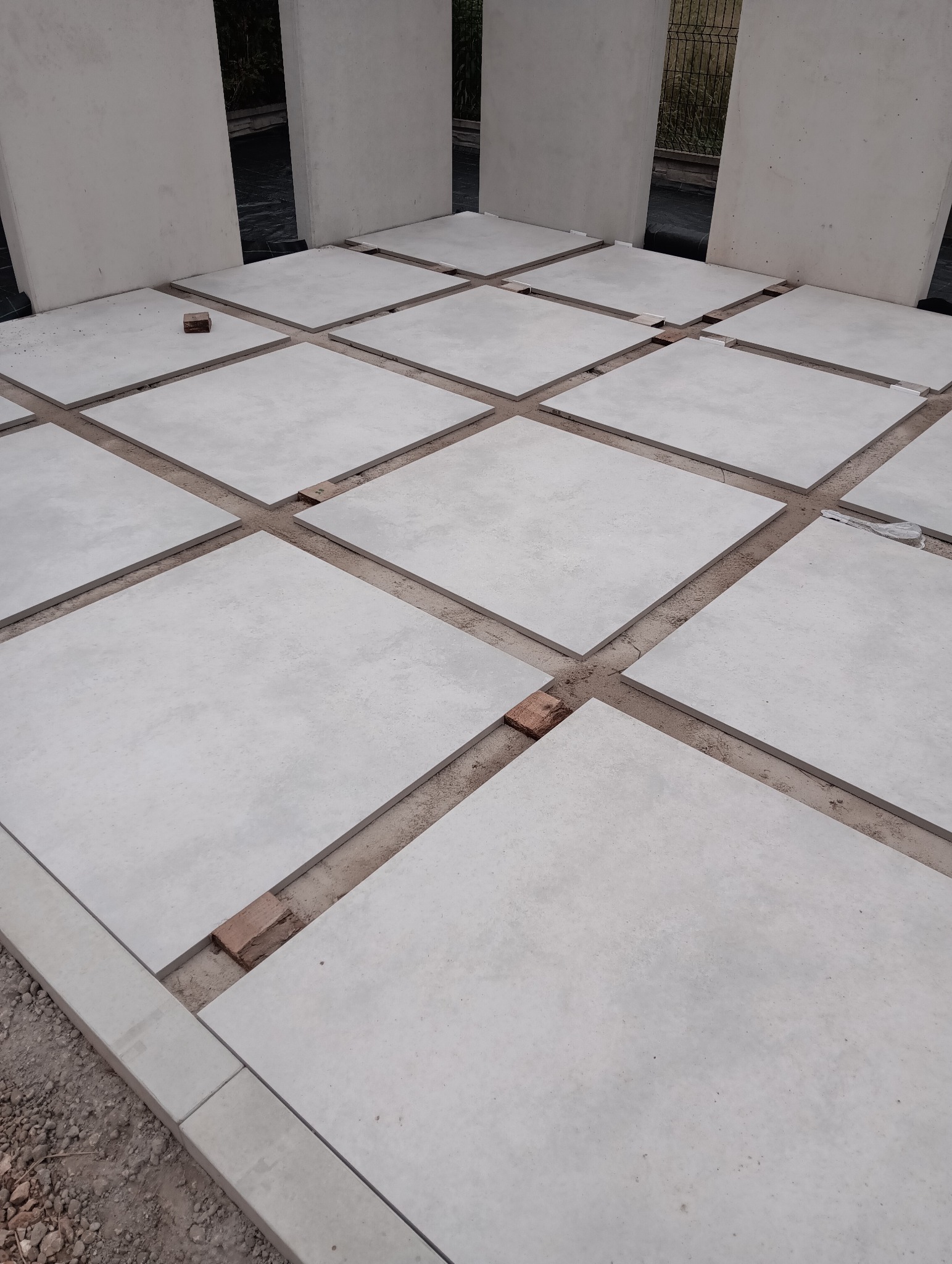 Montaż jasnych, kwadratowych płyt chodnikowych z użyciem cegieł dystansowych, widoczne pionowe elementy ogrodzenia.