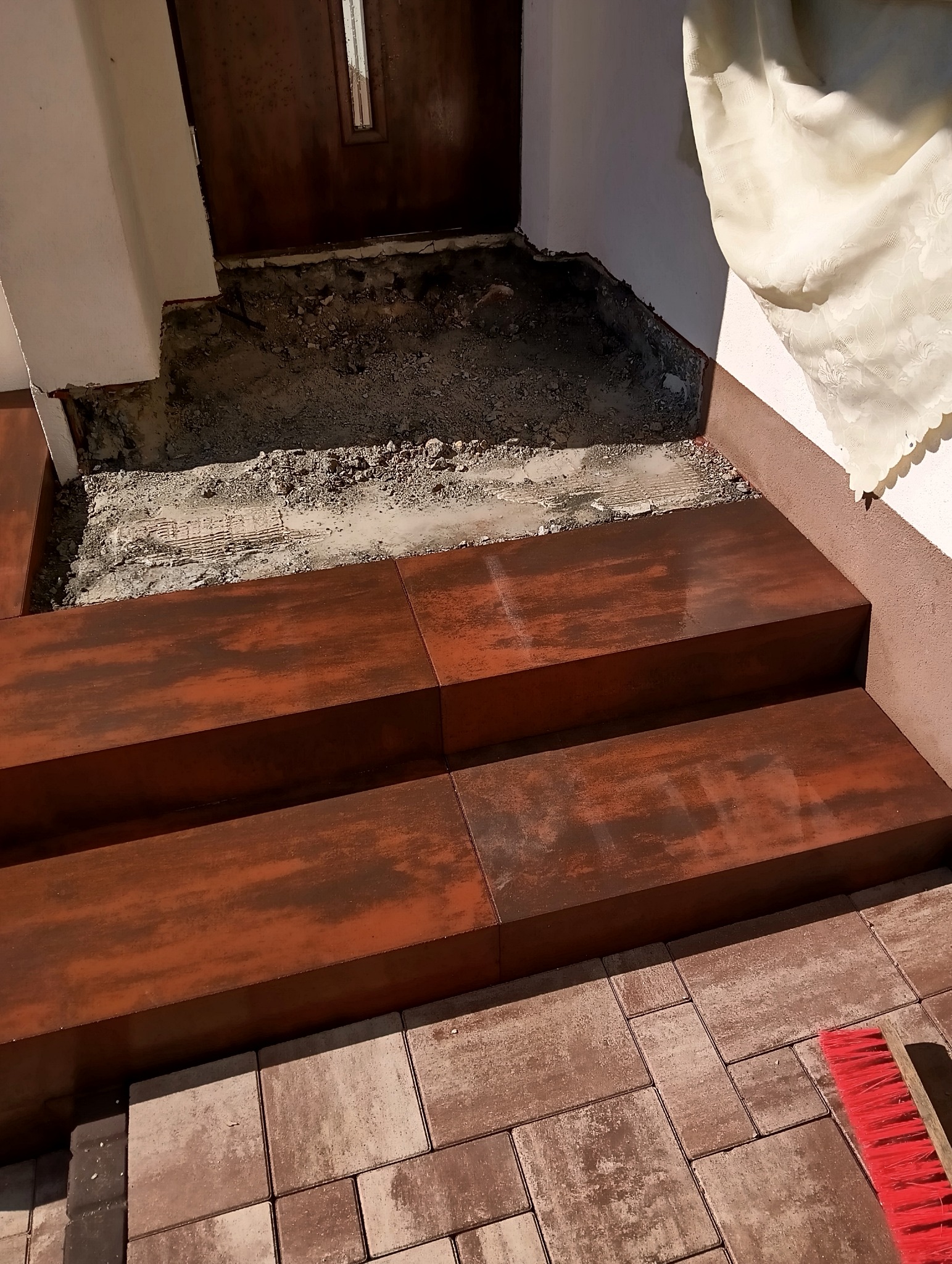 Montaż schodów wejściowych z kostki brukowej o rdzawym odcieniu, widoczny fragment chodnika z kostki i czerwona szczotka. Częściowo wykonane prace.