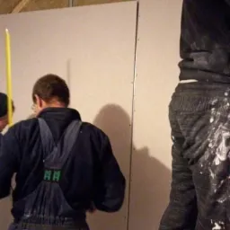 Dwóch mężczyzn w roboczych ubraniach montuje płytę na metalowym stelażu, widoczne ślady farby na ubraniach, jeden stoi na drabinie.