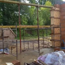 Budowa ściany z cegły z drewnianym szalunkiem, widoczne rusztowanie, deski, wiertarka i rękawice robocze.