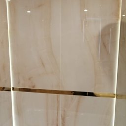 New home kompleksowe remonty i wykończenia wnętrz pod klucz - Beżowe płytki imitujące marmur z dekoracyjnym złotym paskiem i deszczownicą w wykończonej łazience we Wrocławiu.