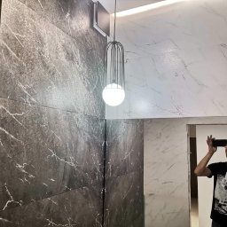 New home kompleksowe remonty i wykończenia wnętrz pod klucz - Wykończona ściana łazienki z ciemnoszarych i białych płytek imitujących marmur, oświetlona wiszącą lampą z metalowym kloszem i kulistym źródłem światła; odbicie fotografa w lustrze.