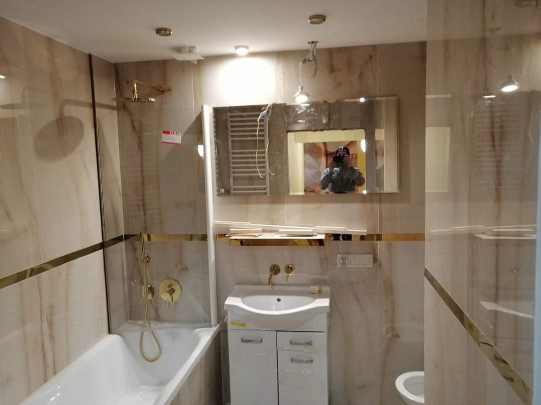 Wnętrze łazienki w trakcie prac wykończeniowych, widoczna wanna z prysznicem, umywalka z szafką, lustro z odbiciem fotografa, marmurowe płytki z złotymi akcentami, częściowo zamontowana armatura.