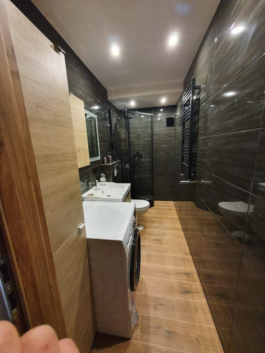 Wnętrze wykończonej łazienki z ciemnymi, błyszczącymi płytkami na ścianach, drewnopodobną podłogą, kabiną prysznicową z czarnymi ramami, białą pralką i umywalką z szafką, oraz grzejnikiem drabinkowym.