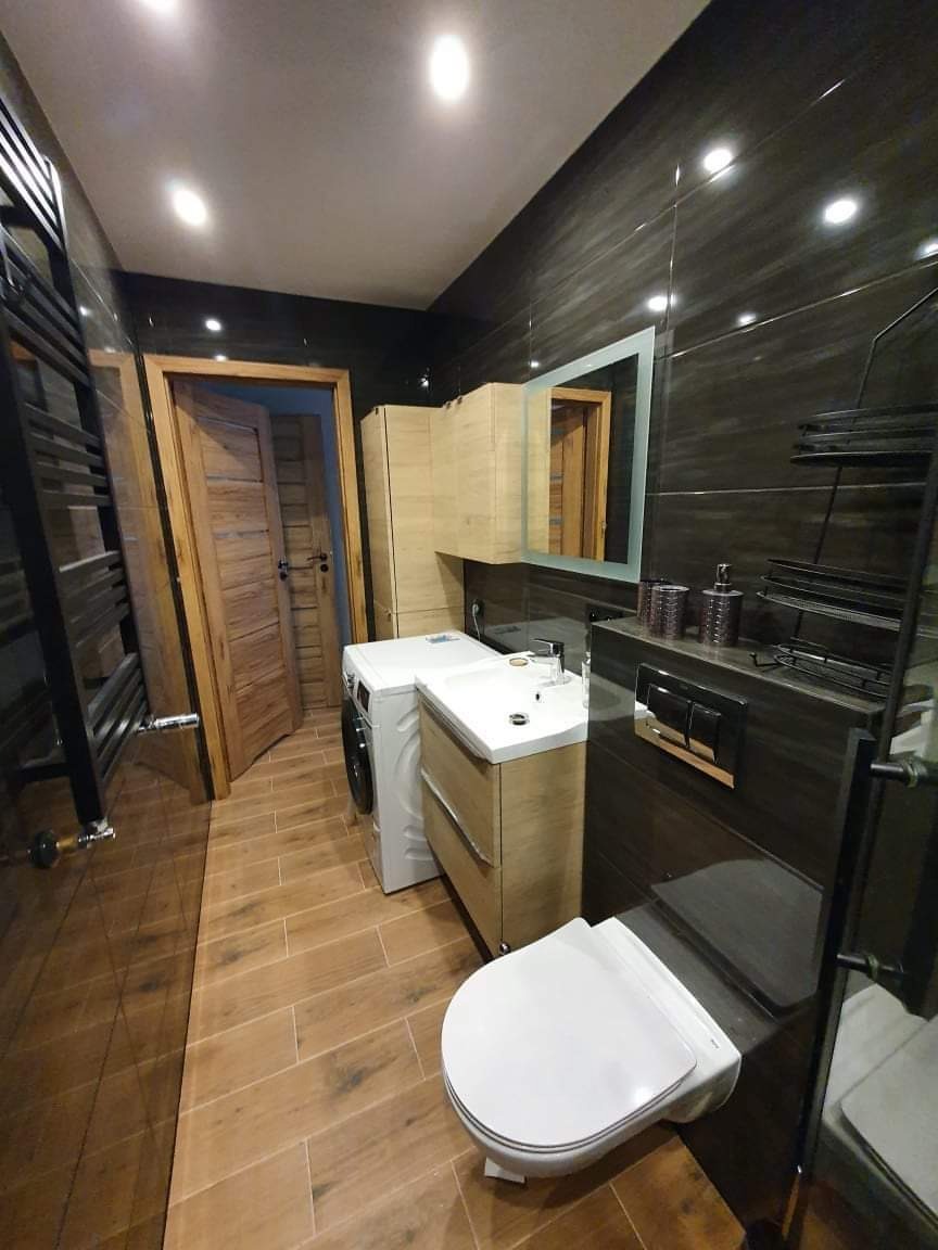 Wykończona łazienka w ciemnych płytkach z drewnopodobną podłogą, wisząca toaleta, pralka obok szafki z umywalką, drewniane drzwi uchylone w tle.