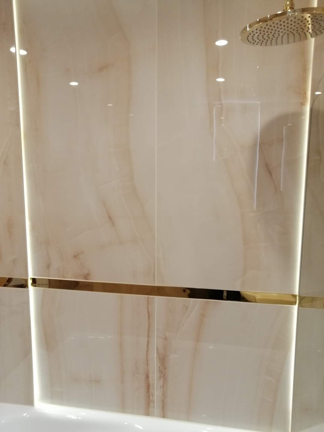 Beżowe płytki imitujące marmur z dekoracyjnym złotym paskiem i deszczownicą w wykończonej łazience we Wrocławiu.