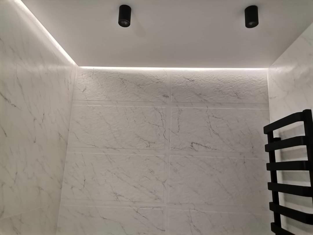 Wykończona łazienka z marmurowymi płytkami, podświetleniem LED wzdłuż sufitu i czarnym grzejnikiem drabinkowym.