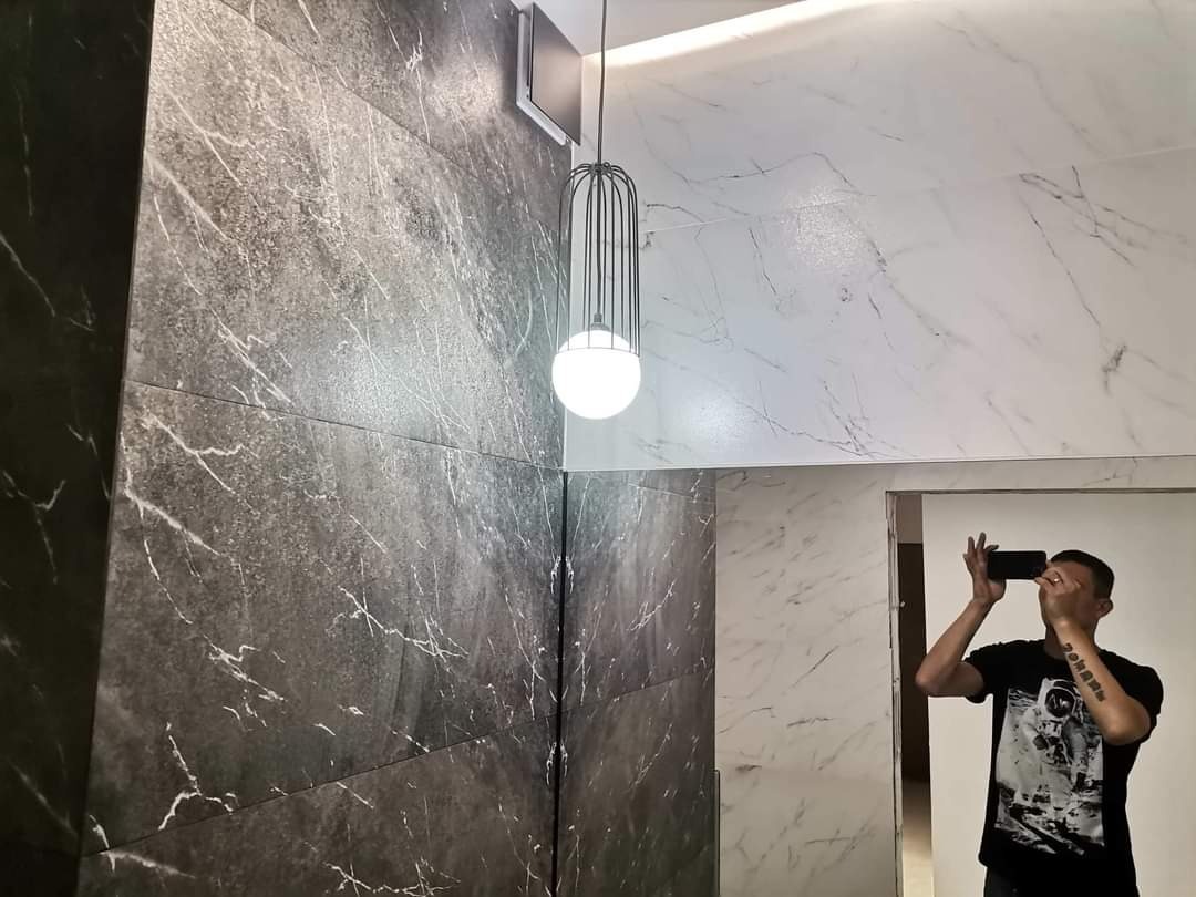 Wykończona ściana łazienki z ciemnoszarych i białych płytek imitujących marmur, oświetlona wiszącą lampą z metalowym kloszem i kulistym źródłem światła; odbicie fotografa w lustrze.