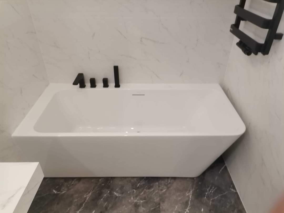 Nowoczesna, prostokątna wanna z czarną armaturą i grzejnikiem drabinkowym na tle marmurowych płytek ściennych i podłogi z ciemnego marmuru.