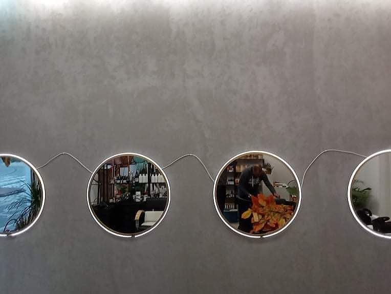 Dekoracyjna ściana z szarą gładzią i czterema okrągłymi lustrami z podświetleniem LED, połączonymi falującym przewodem. W lustrach odbijają się różne elementy wnętrza, w tym osoba pracująca...