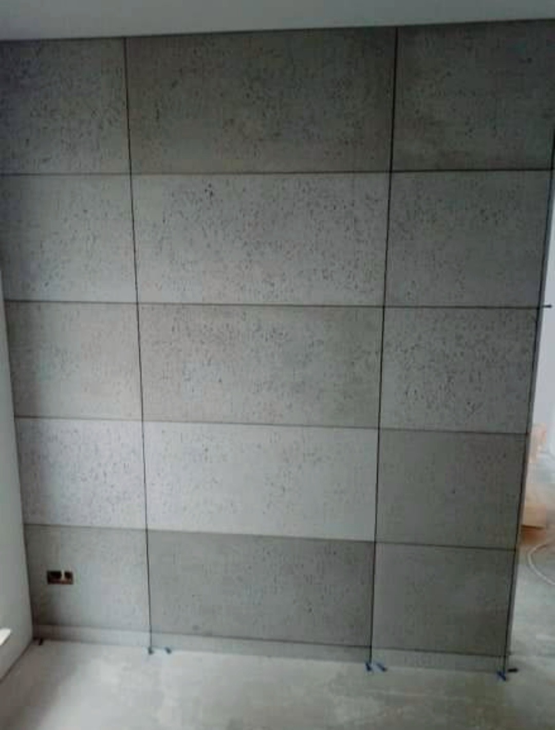 Ściana wyłożona panelami imitującymi beton, z widocznymi fugami i drobnymi dystansami na podłodze, w surowym wnętrzu.