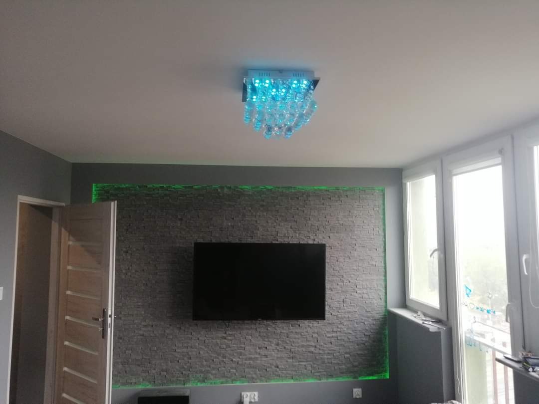 Szara ściana z kamienia dekoracyjnego podświetlona zielonym LED, wiszący telewizor, nowoczesny żyrandol z niebieskim podświetleniem, jasne okno, szare ściany.