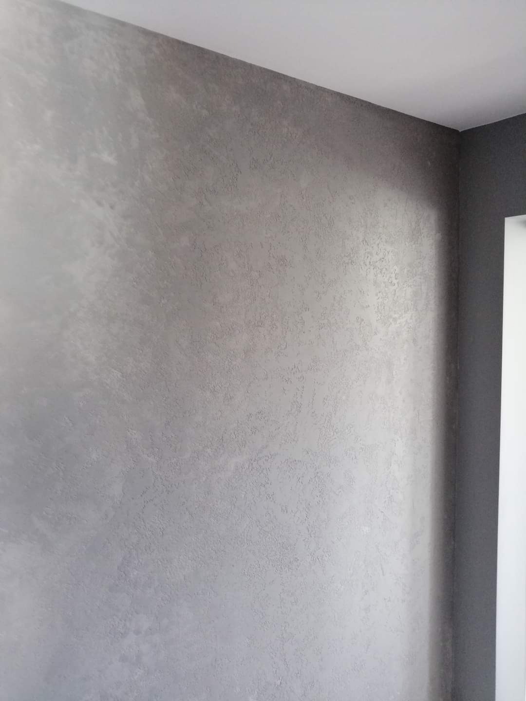 Ściana pokryta dekoracyjną, szarą strukturą, widoczny narożnik pomieszczenia z elementami wykończenia w odcieniach szarości.